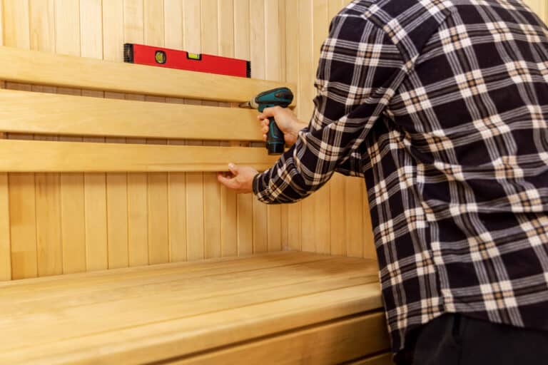 sauna construction - man screwing wooden bench backrest on the wall
