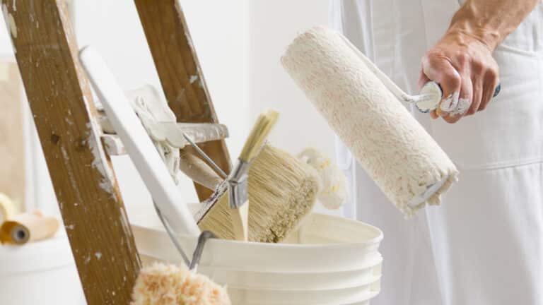 hands of house painter man decorator work of home to renovate, using roller paint and holding white bucket, a wooden ladder with paint brushes as background, close-up